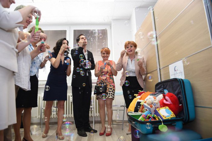 Ilustračný obrázok k článku Pri otvorení centra pokrstili bublinkami kufor plný vyrobených hračiek. Kufor je symbolom cestovania za rodinami, ktoré potrebujú pomoc